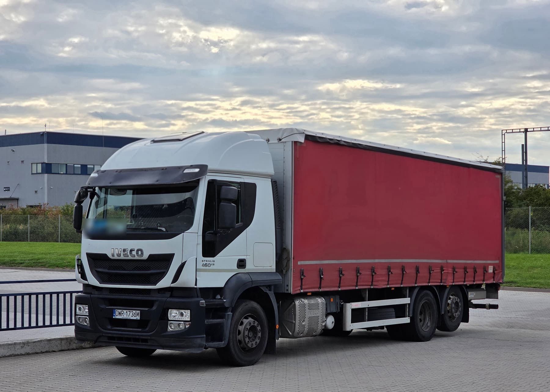 Ciężarówka Markoni: białe Iveco Stralis z czerwoną plandeką, trzy osie, na placu przed magazynem